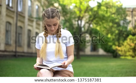 Beautiful teenager girl reading book on bench in campus, preparing homework