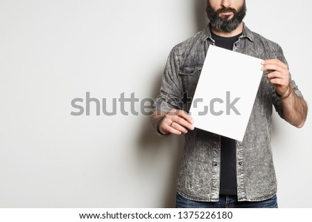 Handsome casual man in a denim shirt shows a white sheet of paper in the camera on a empty background. Mockup Copy Paste Advertisement