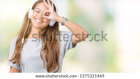 Young beautiful woman wearing headphones listening to music over isolated background doing ok gesture with hand smiling, eye looking through fingers with happy face.