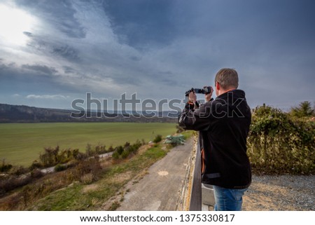 The man takes the camera. A blond man in a black jacket and jeans holds a video camera and takes pictures. The guy is standing over a green valley against a gray stormy sky.