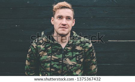 The man is blond on a black wooden background. Guy dressed in a hoodie, camouflage colors
