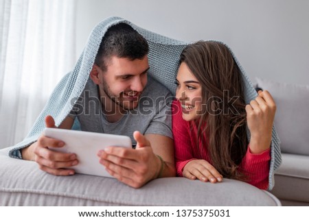 A happy couple are lying in bed under covers while watching a romantic film on the tablet. Adorable young relaxed couple lying under blanket on their bad and reading news on tablet