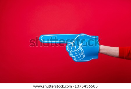 Handsome fan hand glove with foam finge, pointing away over red wall background  Royalty-Free Stock Photo #1375436585