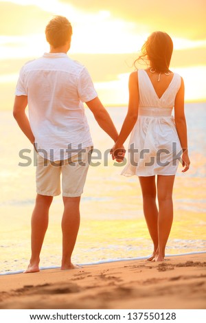 Young couple holding hands at beach sunset enjoying romance and sun. Young happy couple in love on romantic summer holidays vacation. Young lovers in casual clothing. Asia woman, Caucasian man.