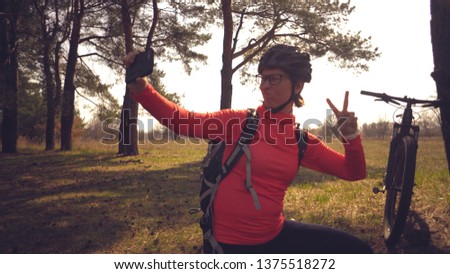 Young caucasian woman athlete tourist cyclist uses hand smart phone photo of herself selfie sitting near tree in coniferous forest outside the city. Sportswoman taking selfie with her mountain bike.