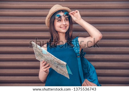 Teenager girl wearing hat carrying backpack standing isolated on wall taking off sunglasses holding map looking aside smiling cheerful