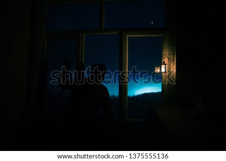 Silhouette of a man looking a dreamlike galaxy through a window. Fantasy picture with old vintage lantern at the window inside dark room.