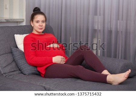 Photo of pregnant woman wears red sweater and maroon leggins with bunch on hand, keeps hand on belly, models looks at camera, sits on gray sofa in living room, has calm expressions. Mathernity concept
