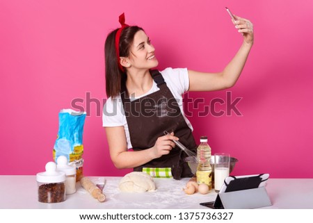 Indoor shot of standing smilling charismatic young lady making selfie at kitchen while cooking new delicious dish, posting photos and videos at social networking sites. Baking and cooking concept.
