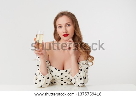 Young beautiful lady with long blond hair, with red lips, holding a champagne glass, thoughtfully looks away cannot decide which dress is still better to buy. Isolated over white background.