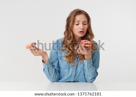 Photo of young beautiful woman with long blond wavy hair, wearing a denim shirt, holds donuts in her hands, gonna bite one of them. Isolated over white background.