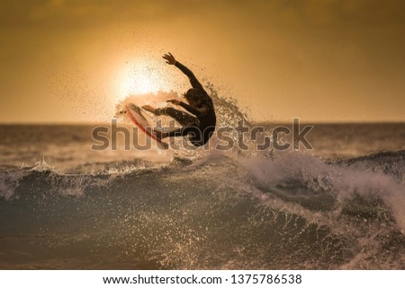 Surfer jumping on the wave at the golden hour on a tropical beach Sporty young man making tricks over the waves with a surf board Extreme sport and action in the water at sunset with foam and spray Royalty-Free Stock Photo #1375786538