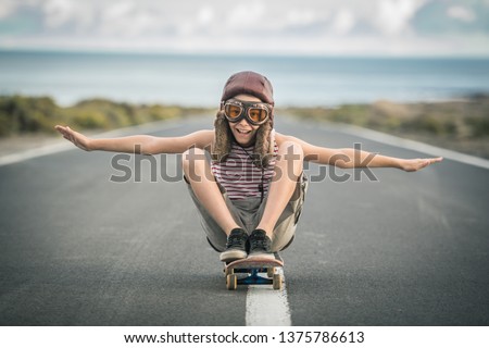 Young blond boy plays aviator sitting cross-legged on skateboard with arms outstretched to fly smiling child imitates plane flying on airport runway. Concept image of a plane taking off for a vacation