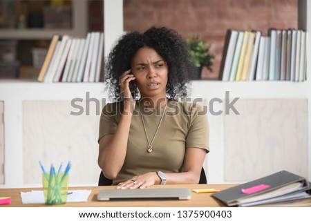 Young serious mixed race office worker female sitting at desk at workplace holding mobile phone makes business call listens client claims feels displeased annoyed and anxious, problems at work concept Royalty-Free Stock Photo #1375900403