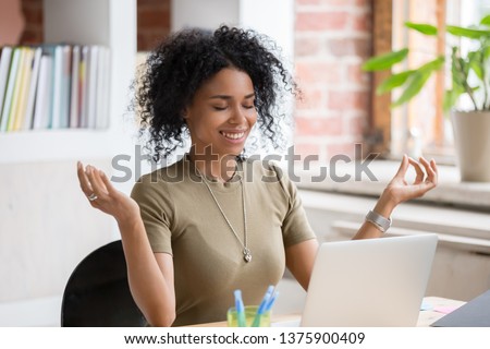 African young woman sitting at table opposite laptop in workplace meditating doing yoga exercise smiling feels good. Take a break, inner balance and harmony, stress relief no negative emotions concept Royalty-Free Stock Photo #1375900409