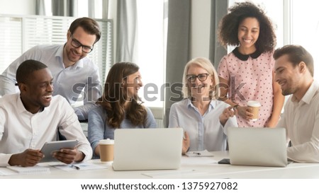 Diverse workers have fun watch video joke online use pc drink coffee break sitting in office room workplace, different ages ethnicity employees members laughing at training with mature business coach Royalty-Free Stock Photo #1375927082