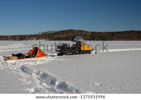 Snowmobile with a sled packed with fishing gear standing on the ice on a lake with forest and mountain in background against a blue sky, picture from Northern Sweden.