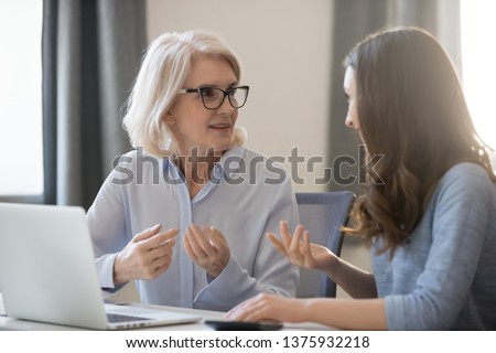 Mature and young women colleagues sitting at desk talking about project startup ideas, sharing thoughts, solve currents issues, make research, discuss growth strategy, think how generate more revenue Royalty-Free Stock Photo #1375932218