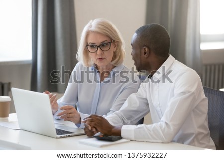 Serious experienced aged businesswoman in glasses explains help to african new employee task usage of corporate online program sitting at table office room working together, mentor and trainee concept Royalty-Free Stock Photo #1375932227