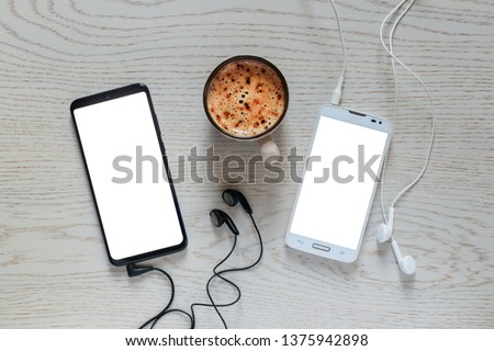 Two smartphones with connected earphones and isolated blank white screens for design on the background of a desk with a cup of coffee