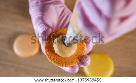 Pastry chef is filling cream lemon macaroons with pastry bag. Hands in gloves close-up. top view