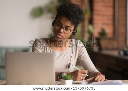 Focused mixed race woman wearing headphones watching webinar write notes study online with online teacher, african female student learning language computer course on laptop listen translate lecture