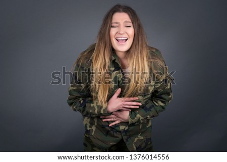 Joyful European female soldier keeps hands crossed, laughs at good joke, wears casual clothes and round spectacles, standing indoors. Happy young woman with long hair poses against gray wall.