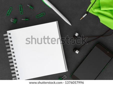 Mobile phone with blank white spiral paper notebook, pen, paper clips, paper clumps, push pins, headphones and green cloth on empty dark background top view
