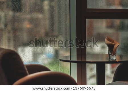 Design interior with coffee table and armchair near the window. Raindrops on the window glass. Blurred cityscape on background. Overcast rainy weather. Selective focus on window.  Royalty-Free Stock Photo #1376096675