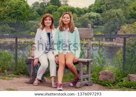 Mother and daughter posing for a family picture. Adult women sitting on a wooden bench near the river in a country side.