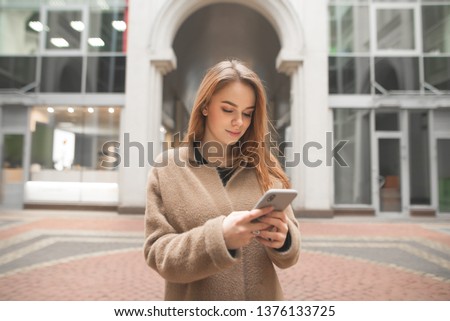 Attractive stylish woman in a coat,stands on the background of modern architecture and uses a smartphone.Urban portrait of a beautiful woman with a smartphone in hand,looking at the screen and smiling