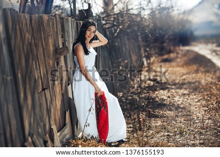 Portrait of a Beautiful black haired girl in a white vintage dress standing near wood fence.Young woman model posing in a russian national style . red shawl.