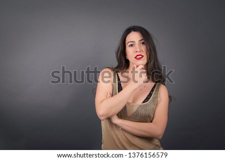 
Portrait of thoughtful smiling girl keeps hand under chin, looks directly at camera, listens something with interest, dressed casually, poses against gray wall. Youth concept.
