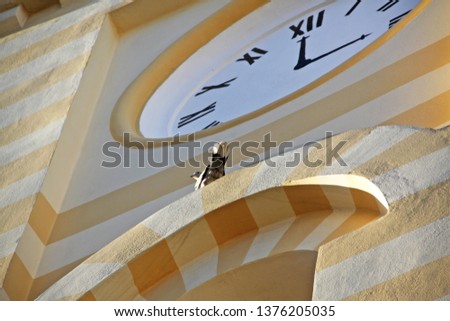 Street clock on the tower. Yellow tower. Pigeon sitting on the structure. Bottom-up view.