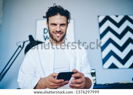 Portrait of smiling caucasian millennial man holding mobile phone connected to wifi in office for chatting, handsome businessman satisfied with modern gadget and apps for search information
