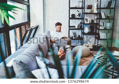 Serious caucasian man using laptop computer for paying and shopping online via wifi, hipster guy lying on sofa in living room with netbook watching videos during free time at home interior Royalty-Free Stock Photo #1376298674