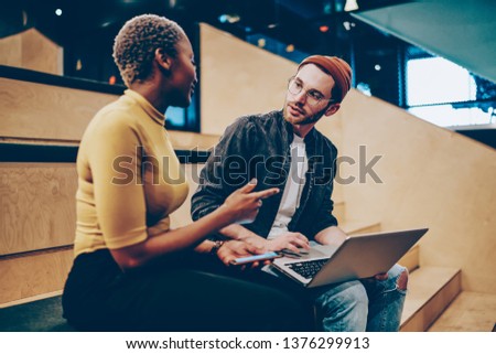 Young male and female colleagues having conversation about business sitting together with laptop computer, trendy dressed hipster guy listening attentively to female speak about her creative ideas
