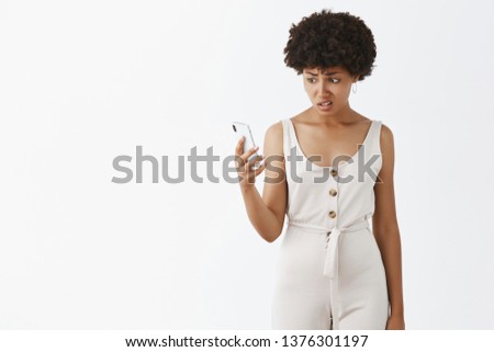 Woman cannot udnerstand who called from unknown number and told rude things. Confused clueless and intense cute african american in white stylish overalls staring questioned at smartphone screen