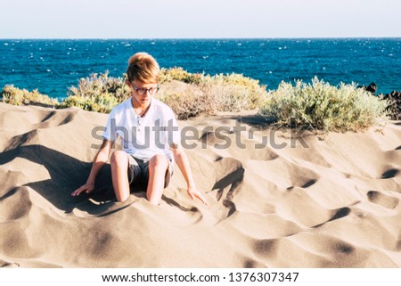 young boy playing alone with sand at the beach - traveler and vacation lifestyle, caucasian child with glasses sit - sea at the background