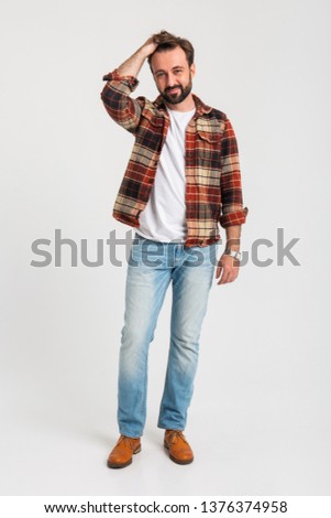 isolated handsome bearded man in hipster outfit dressed in jeans and checkered shirt full length standing on white studio background fixing his hair, attractive guy