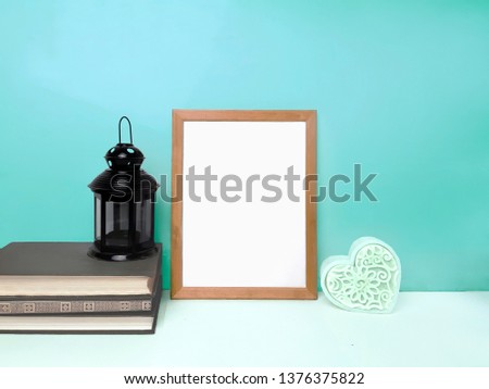 Blank frame mockup, decorative heart, stack of books, napkinlantern on table over mint colored wall. Minimalistic design
