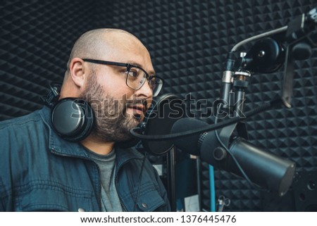 Young fat and bald radio presenter or host or dj in glasses  with headphones around his neck speaks into microphone at radio studio, close up portrait