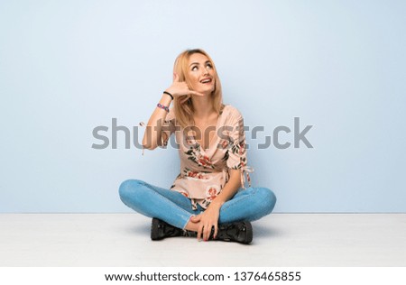 Young blonde woman sitting on the floor making phone gesture. Call me back sign