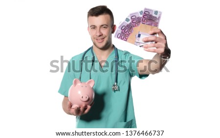 Smiling male doctor holding piggy bank and stacks of money isolated on white background 