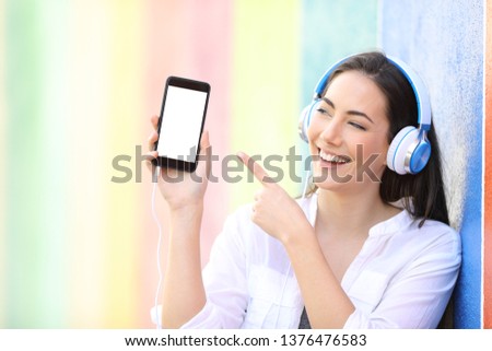 Happy girl listening to music showing and pointing smart phone blank screen in a colorful background