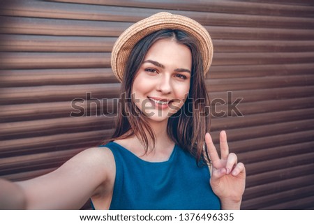 Teenager girl wearing hat standing isolated on wall taking selfie pictures showing peace sign looking camera smiling cheerful face close-up