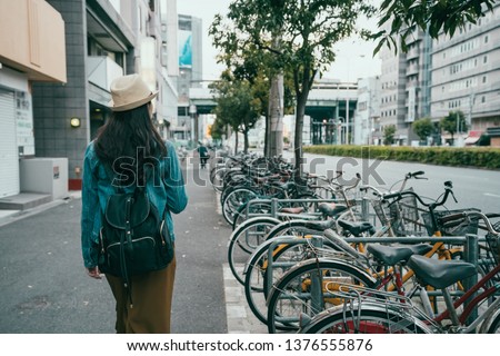 Back rear view of hipster girl walking on city street in osaka japan. young female backpacker in hat go through bikes in parking lot for bicycles in modern urban walkway with shop close on side.
