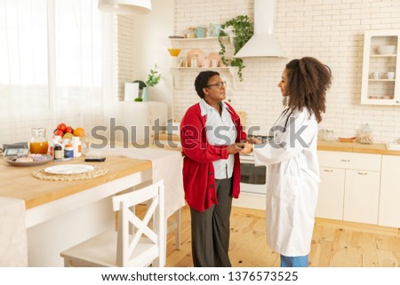 Pleasant nurse. Aged woman wearing red cardigan feeling grateful to pleasant nurse taking care of her