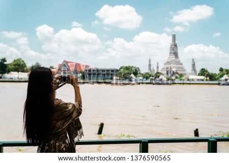 Back view of Asian woman taking photo of Buddhist temple by mirrorless camera from riverside.