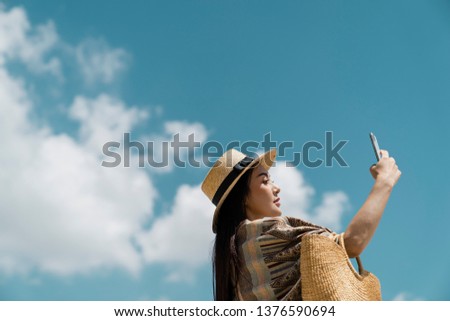 Side View Portrait of Attractive Asian traveler wearing traditional clothes , brown boater,carrying straw bag taking photo by smartphone with fresh blue sky background.